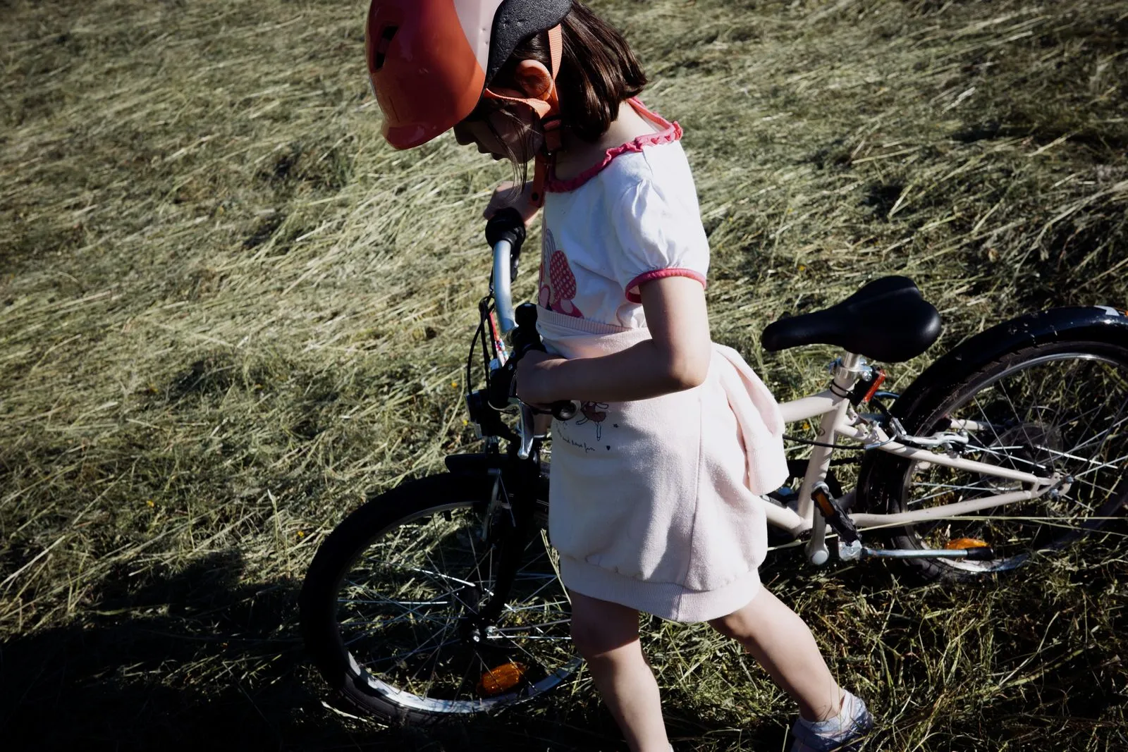 Young girl wearing helmet walking her bicycle through a grassy field on a sunny day.